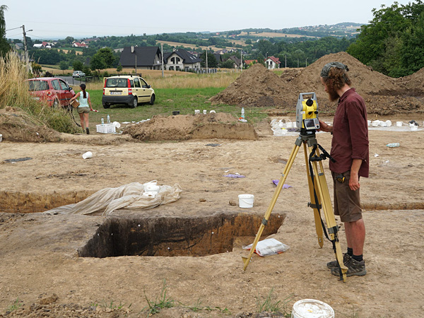 A man is pointing a geodetic instrument at a dug hole. He protects his head from the sun with a scarf