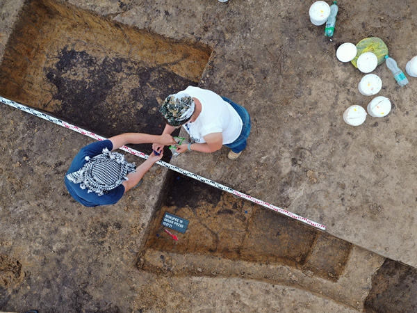 View from above of two dug holes with dark colored bottoms. Two women wearing headscarves are standing at their sides