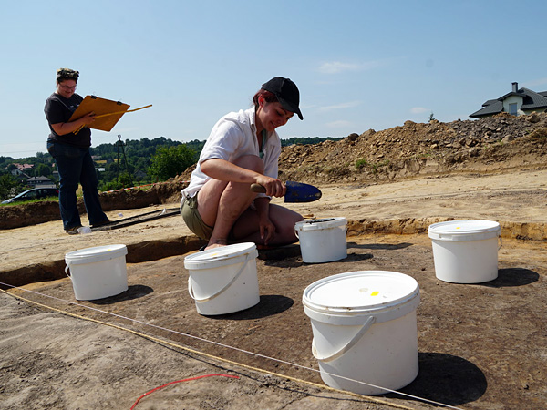 A woman in a hole uses a shovel to collect samples into plastic buckets. Another woman stands behind her with a sketchbook