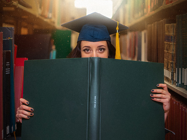 A woman in a student cap is standing between library shelves. She is looking over the cover of her thesis