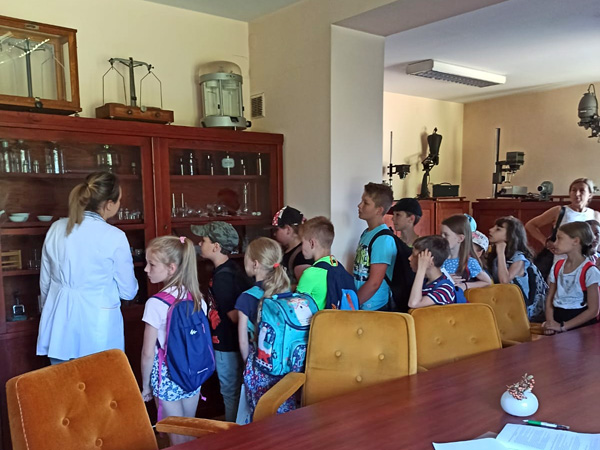 A woman in a white coat stands among school children. They all stare at the contents of a glass display case