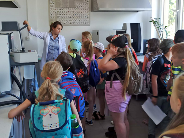A woman in a white coat stands among school children in a modern laboratory