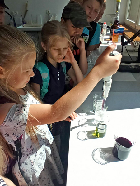 A girl uses a pipette to inject purple liquid into a glass beaker of green liquid. Other pupils watch her