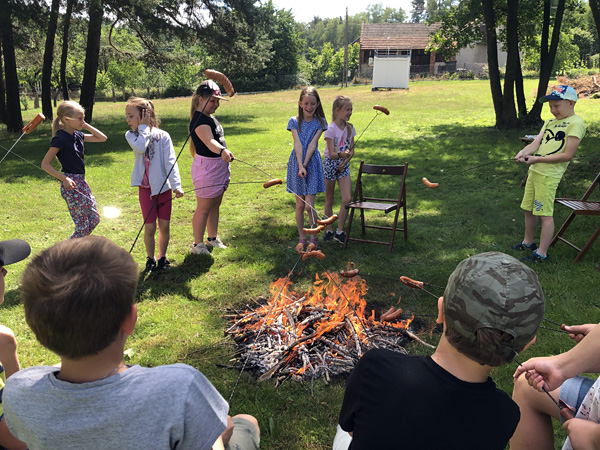 Children stand around a campfire. They hold sausages on skewers above it