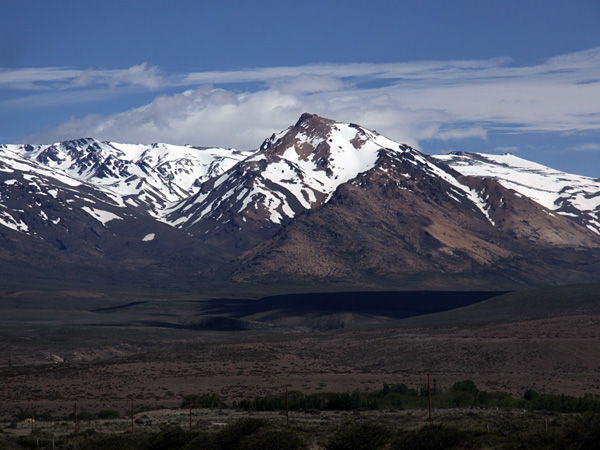 A chain of mighty mountains with snow-capped peaks. At its foot is a vast, almost flat area, sparsely covered with vegetation