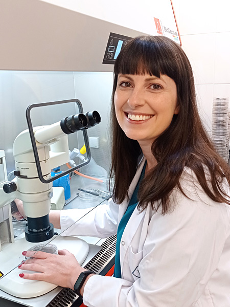Smiling dark-haired woman in lab coat sits at microscope