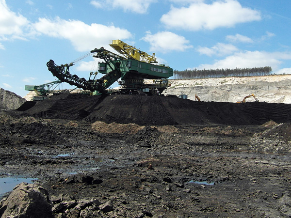 A huge mining machine stands on a black pile of lignite. In the distance, the light yellow, almost white walls of the open pit