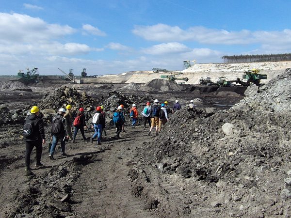 A group of people in hard hats walk across dark, rocky ground. In the distance, huge mining machines