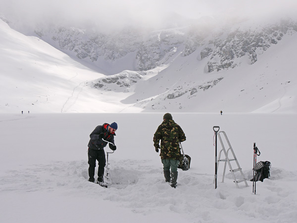 Two men stand on the frozen, snow-covered surface of a pond. One of them is drilling a hole in the ice with a large auger