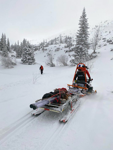 A snowmobile pulls a loaded sledge. In the distance, a person with trekking poles walks through the snow among sparse spruces