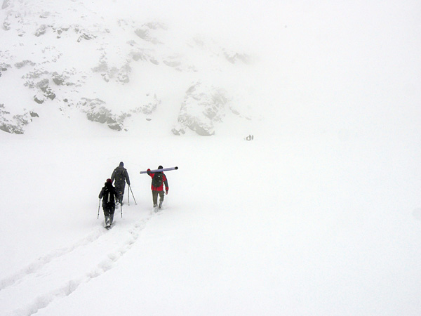Three people in an completely white, snowy landscape. They leave a trail in the deep snow. One is carrying a long object