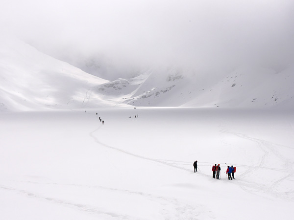 A distant view of a group of people standing on a frozen and snow-covered pond surrounded by white mountain slopes