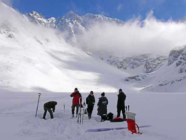 Several people set up their equipment on the surface of a frozen pond. The pond is surrounded by snow-capped mountains