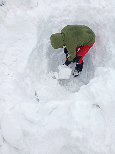 View from above of a person digging a hole with a shovel in snow. He is wearing winter hiking clothes and a hood on his head