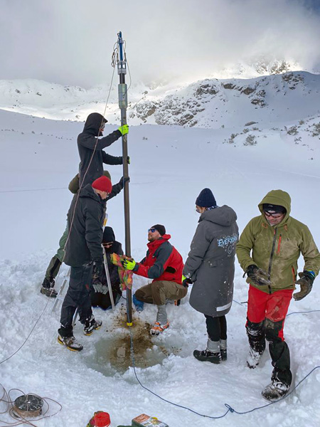 Several people at a hole dug in the snow and ice. Some of them are holding a long tube-shaped device vertically