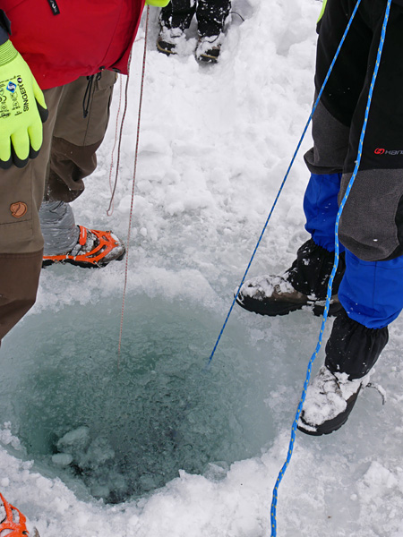 Two people, visible from the waist down, stand by a hole in the ice. They hold thin ropes that sink into the water