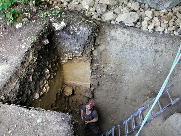 A woman in a hard hat stands at the bottom of a deep pit with straight, vertical walls. A ladder leans against the pit wall