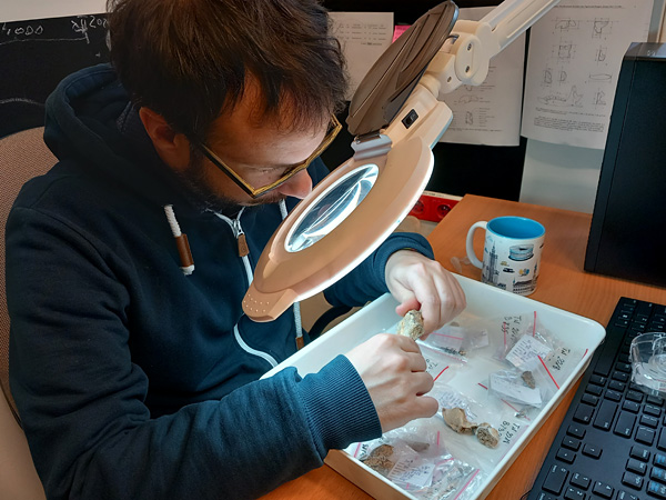 A man sits at a desk and looks at a small animal skull through a large magnifying glass mounted on a swing arm