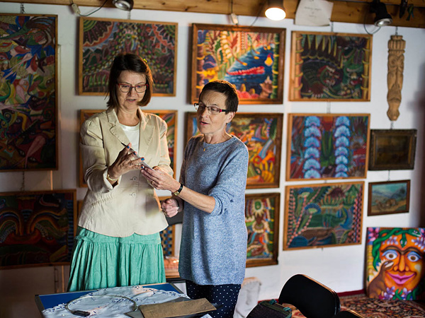 Two women stand in a gallery in front of a wall hung with paintings of colorful, fantastic landscapes