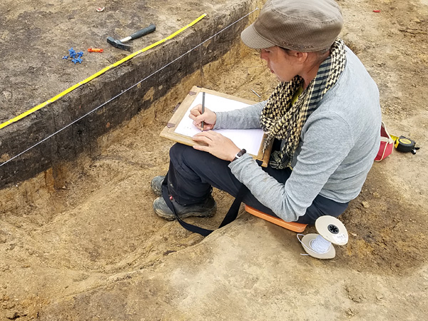 A woman sits on the edge of a shallow hole, sketching something on a white sheet of paper. She looks at the dark soil profile