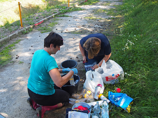 Two women kneel on a path. One of them is writing on a plastic bag with a marker. There are buckets and bottles nearby