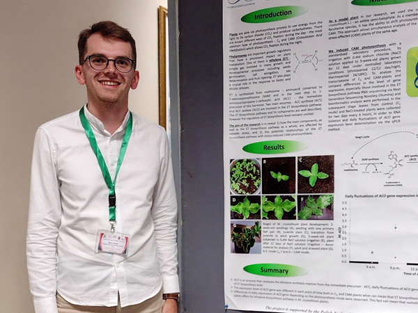 A young, smiling man in a white shirt and with an ID tag around his neck stands next to a poster presenting his research