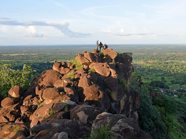 A brown rock towering over an endless green plain in the light of the afternoon sun. Three people stand on its top