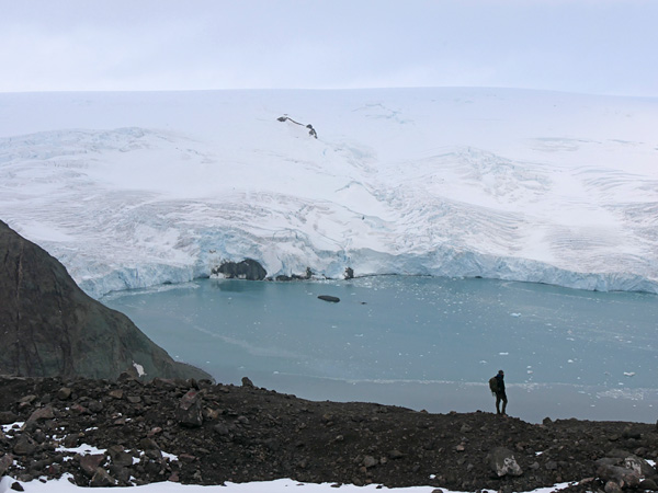 A black rocky shore with a small silhouette of a man. In the background, a frozen surface of water and an endless glacier