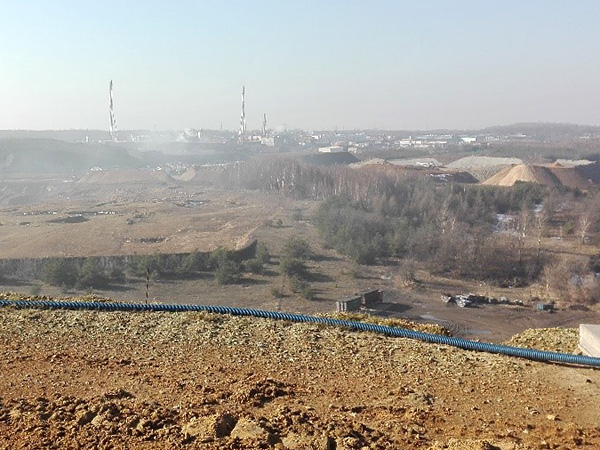 View from a high mound onto an industrial landscape with heaps of waste and tall chimneys, barely visible in the dusty air