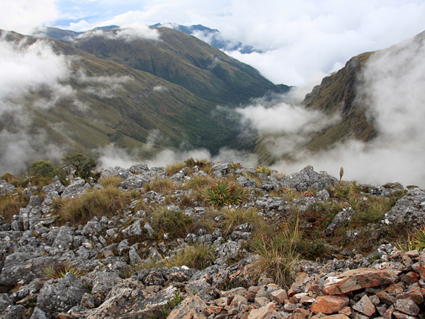 A view from above of partially cloud-covered mountains and a green valley. In the foreground, sparse grass on a stony slope
