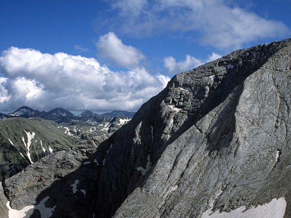 A grey, almost smooth rock wall, partially obscuring the view of the mountain range. There is some snow in rock crevices