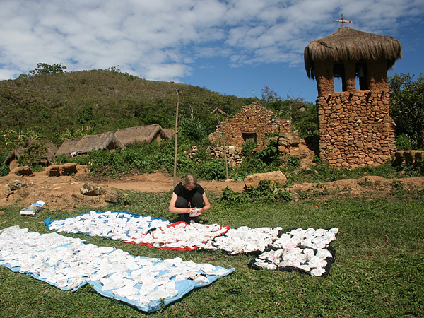 A woman crouches by the tarpaulins with numerous white bags stacked on them. In the background, a primitive mountain village