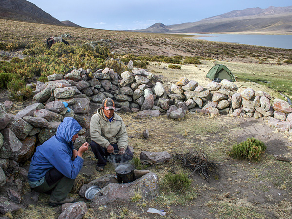 Two people crouch by a steaming cauldron. They are sheltered by a stone wall. In the background, a tent and a mountain lake