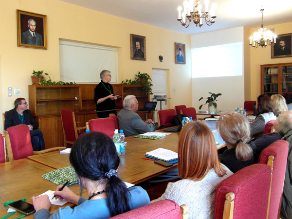 People are sitting at a long wooden table, looking at a presentation screen. On the yellow wall behind them hang portraits