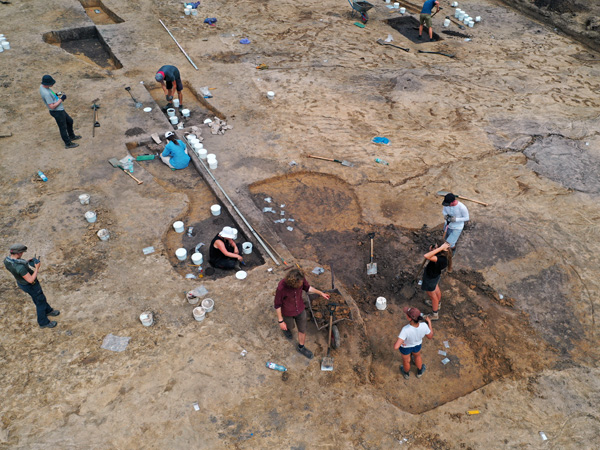 A view from above of a group of people working on several shallow holes. Plastic buckets are spread out on the ground nearby