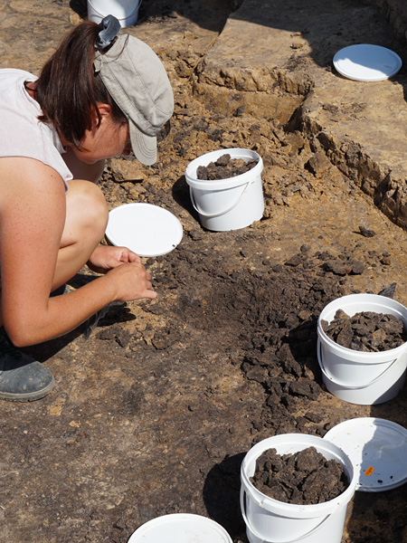 A woman crouches in a shallow hole. Next to her are white plastic buckets filled with clods of earth