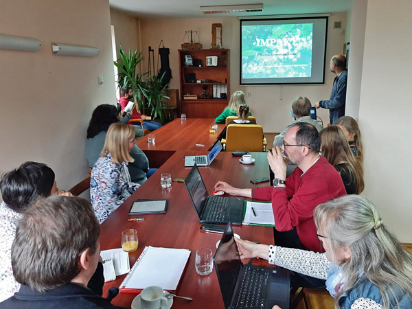 A group of people sit at a long table with laptops, notebooks and cups. In the background, a man displays a slide on the wall