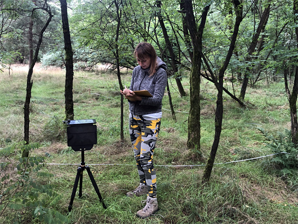 A woman dressed in sportswear stands in a forest next to a device mounted on a tripod. She writes something in a notebook