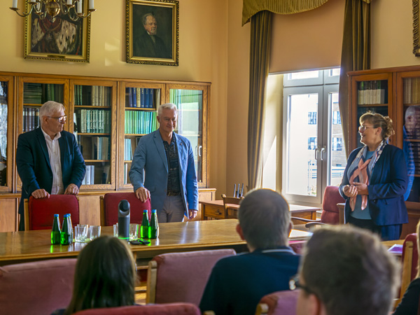 Two men stand behind a large wooden table. A woman stands to the side introducing them to the audience sitting in armchairs