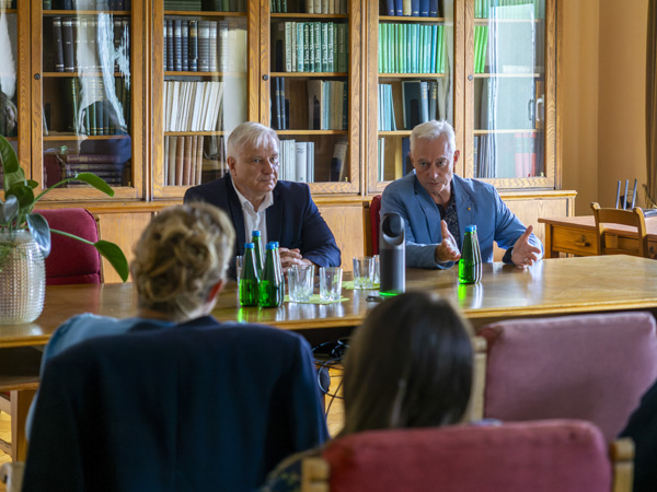 Two men sit behind a large wooden table. One of them gesticulates and speaks to the audience
