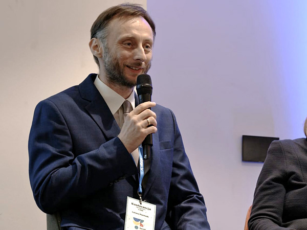A man in a suit, shown from the waist up, speaks into a microphone while smiling at the audience