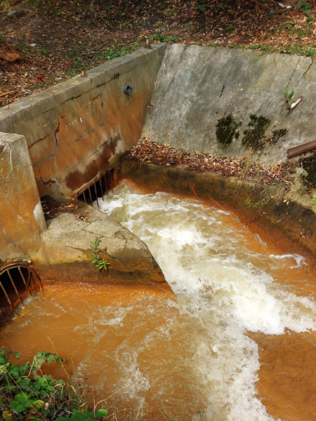 Foamy, rusty water flows from the concrete-walled channel into two holes covered with grates