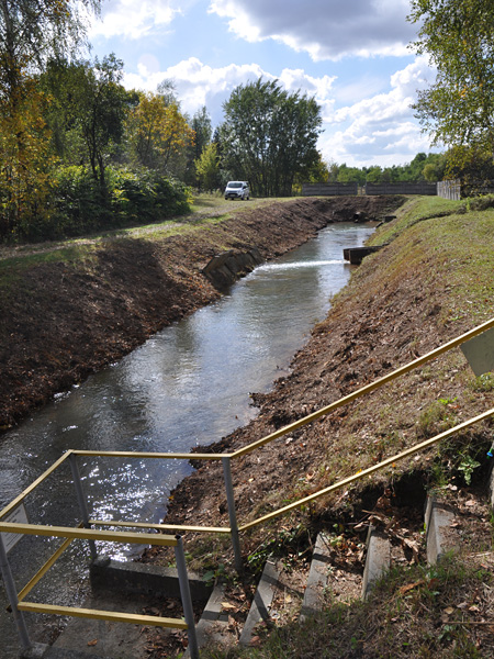 A narrow watercourse with straight, embanked banks. In the foreground, steps to the water. In the background, a parked car