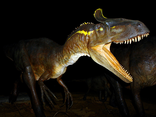 A model of a predatory brown dinosaur emerging from the shadows. It has a wide open mouth and a yellow stripe on its neck