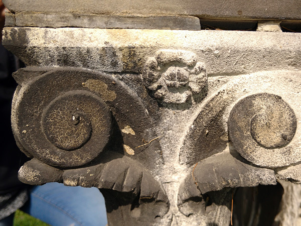 Close-up of the capital of an old, light grey, stone monument, half covered with a dark brown coating